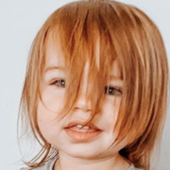 A red-haired toddler in a grey sleeper, hair falling over the eyes